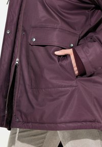 Růžově hnědá nepromokavá bunda s předním zipem, boční kapsou se zacvakávacím knoflíkem a hladkým povrchem. Spárována s béžovými manšestrovými kalhotami.