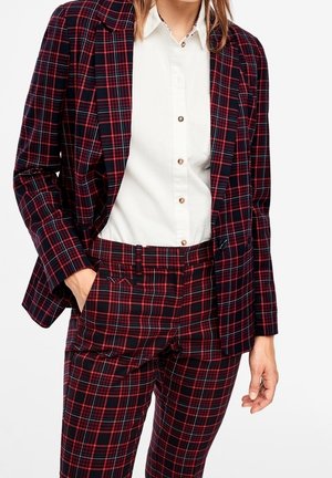 Kobieta ubrana w czerwoną i czarną kratowaną marynarkę oraz spodnie, do kompletu, na białej koszuli zapinanej na guziki, z ręką w kieszeni.