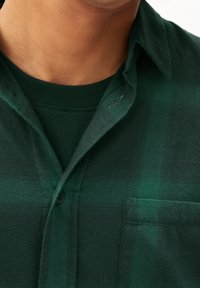 Primo piano di una persona che indossa una camicia a quadri verde scuro sopra una maglietta verde coordinata.