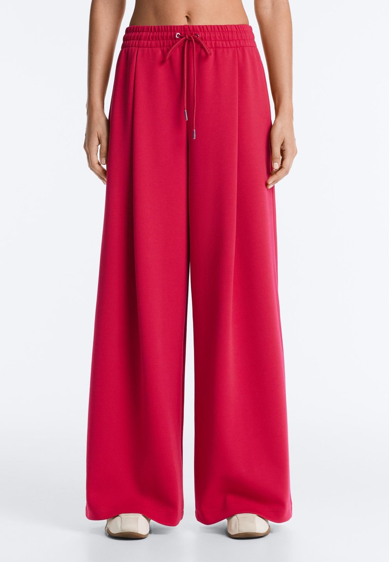 Pantalon rouge à jambes larges avec taille élastique et cordon, porté par une personne avec le ventre nu et des chaussures blanches sur fond blanc.