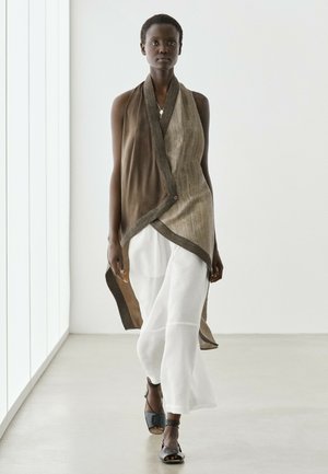 Große Person, die in leichten, luftigen weißen Hosen, asymmetrischem braun-beigem ärmellosem Top und schwarzen offenen High Heels in einem hellen minimalistischen Raum geht.