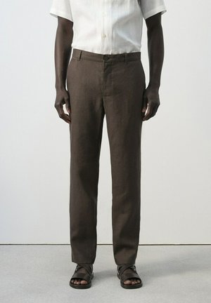 Hombre vistiendo pantalones marrones de pierna recta, una camisa blanca de manga corta abotonada y sandalias marrón oscuro abiertas en los dedos, de pie contra un fondo liso.