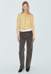 Gul knäpp cardigan över en vit skjorta, kombinerad med mörkbruna raka jeans. Modellen har på sig svarta klackskor. Enkel bakgrund.