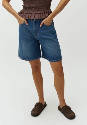 Woman wearing dark blue denim shorts, brown ruched sleeveless top, brown slip-on clogs, standing with hands in pockets.