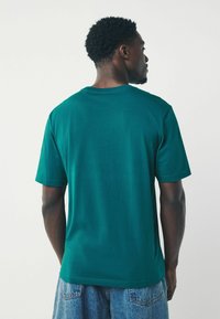 Camiseta de algodón color teal, cuello redondo, mangas cortas, textura suave. Usada con pantalones cortos de mezclilla claros, vista desde la espalda.