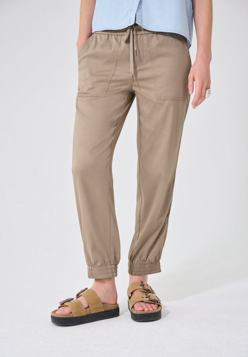Pantalon beige à taille élastique avec cordon de serrage et poches latérales, associé à des sandales marron clair à boucle, porté par une personne debout sur un fond uni.