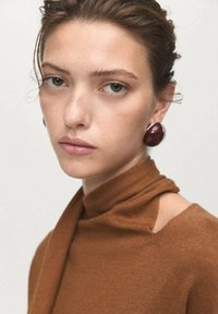 Giovane donna con i capelli raccolti che indossa un maglione marrone a collo alto e grandi orecchini a forma di goccia di colore rosso scuro, sfondo neutro.