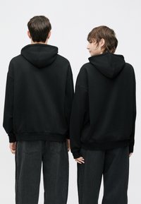 Juodi gobtuvu megztiniai pagaminti iš minkštos medžiagos su laisvu siluetu. Turi didelį gobtuvą ir ribotuotas rankogalius, pavaizduoti dviejų asmenų iš nugaros.