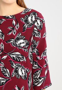 Tissu à motif floral bordeaux avec de grandes fleurs noires et blanches surlignées de rose. Texture douce, manches larges et décolleté bateau.
