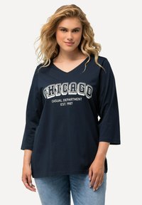 Marineblaues V-Ausschnitt-Shirt mit drei Viertel-Ärmeln, auf dem in fetten weißen Buchstaben "CHICAGO CASUAL DEPARTMENT EST. 1987" gedruckt ist.