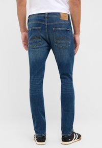 Vīrietis, kurš ģērbjas šaurās zilo džinsu biksēs ar izšūtiem aizmugurējiem kabatām un melnām sportiskajām apaviem ar baltām svītrām, stāv uz baltā fona.