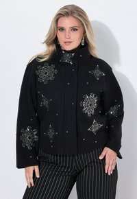 Femme portant une veste noire avec des motifs de flocons de neige en perles argentées et un pantalon noir à rayures, posant sur un fond blanc uni.