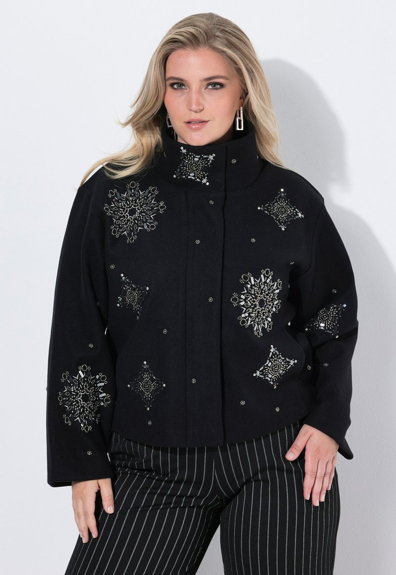 Femme portant une veste noire avec des motifs de flocons de neige en perles argentées et un pantalon noir à rayures, posant sur un fond blanc uni.