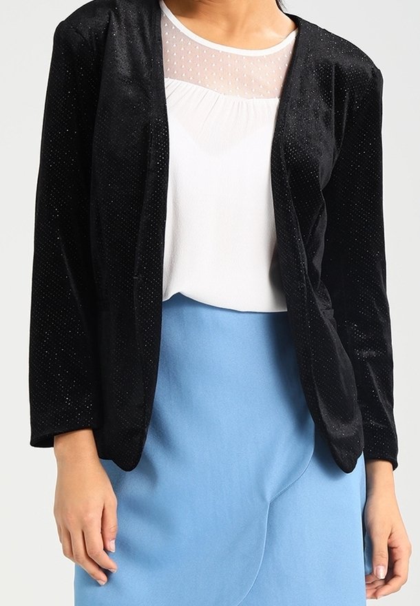 Personne portant un blazer en velours noir, un chemisier blanc avec un col transparent à pois et une jupe bleu clair sur un fond uni.