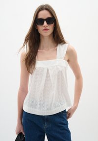 Haut blanc sans manches avec un décolleté carré et un motif texturé. Associé à un jean en denim bleu foncé et accessoirisé avec des lunettes de soleil noires.