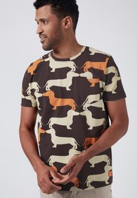 Kortärmad t-shirt i brunt bomull med ett upprepande mönster av taxar i krämfärgat och orange, med en liten bröstficka.