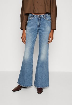 Osoba ubrana w brązową krótką koszulę, jasnoniebieskie spodnie z wysokim stanem i rozszerzanymi nogawkami oraz ciemnobrązowe spiczaste buty na białym tle.