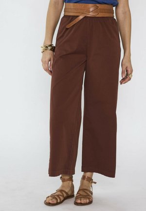 Personne portant un pantalon marron à jambes larges avec une ceinture beige à nouer, des sandales marron à brides, des bracelets dorés et un haut bleu (torse partiellement visible).