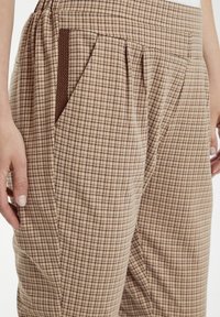 Bruna rutiga byxor med en strukturerad tygkvalitet, sidfickor, veck och en figursydd midja. Mönstret består av små rutor i ljusa och mörka nyanser.
