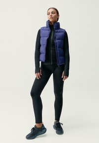 Vijolična oblazinjena brezrokavnik z visokim ovratnikom, oblečen čez črn dolg rokav in črne legice, dopolnjen s temnimi športnimi čevlji.