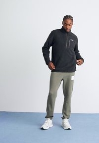 Man with braided hair wearing a black zip-up fleece, gray sweatpants with white patch, and white sneakers, standing on a blue floor.