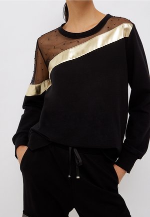 Vrouw draagt een zwart shirt met lange mouwen met een doorschijnende versierde bovenkant en een diagonale metalen gouden streep, gecombineerd met zwarte broek met trekkoord.