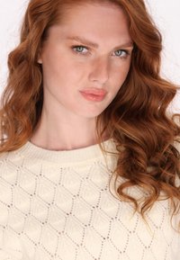 Krém színű kötött pulóver, amelyet gyémánt mintázat és bordás nyakú kialakítás jellemez, kiegészítve laza, hullámos vörös hajjal és természetes sminkkel.