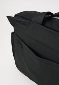 Sort stof messenger tas med en tekstureret overflade, lynlåslukning og lommer sikret med velcro. Inkluderer sideopbevaringsrum for ekstra plads.