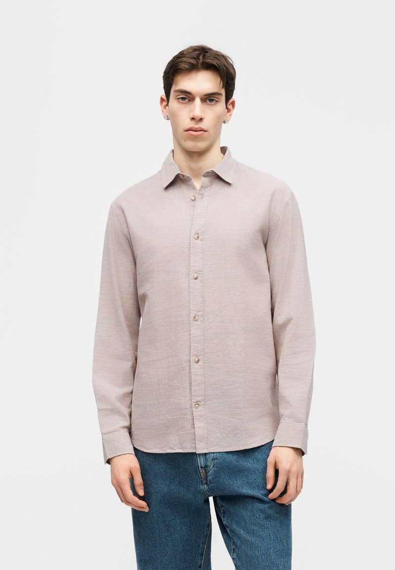 Νέος άνδρας με κοντά σκούρα μαλλιά, που φοράει ανοιχτόμπεζ πουκάμισο με κουμπιά και μπλε τζιν, στέκεται μπροστά σε απλό λευκό φόντο.