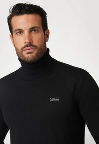 Maglione a collo alto nero realizzato in materiale morbido, con dettagli a coste e un piccolo logo bianco "Guess" sul petto.