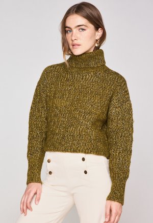 Olivgrüner Strickpullover mit hohem Kragen, taillierter Silhouette, gerippter Textur und langen Ärmeln. Kombiniert mit hellen Hosen mit Knopfdetails.