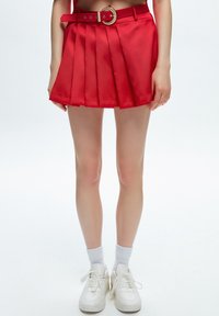 Mini-jupe plissée rouge avec une taille et une boucle de ceinture ronde. Associée à des baskets blanches et des chaussettes mi-mollet. Texture de tissu lisse.