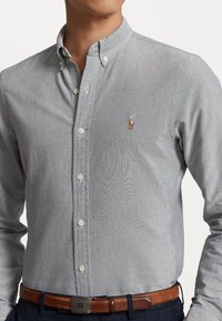 Uomo che indossa una camicia button-down grigio chiaro con un piccolo logo ricamato di un giocatore di polo e una cintura in pelle marrone sopra dei pantaloni scuri.