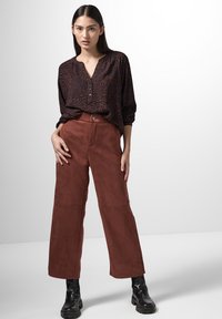 Braune Wildleder-Hose mit weitem Bein und vorderem Knopfverschluss, kombiniert mit einem dunkel gemusterten Hemd mit Knopfleiste, V-Ausschnitt und lässiger Passform.