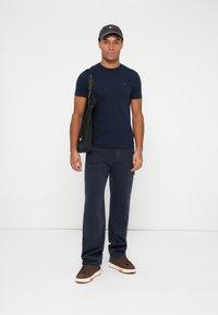 Muž v námorníckom tričku, tmavých nohaviciach, hnedých teniskách a čiernej čiapke stojí s čiernou taškou cez rameno na bielom pozadí.