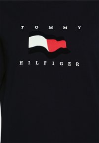 Marineblaues T-Shirt mit einem auffälligen bestickten Flaggenmotiv in Rot, Weiß und Blau. Der Text "TOMMY HILFIGER" in Weiß unter dem Logo.