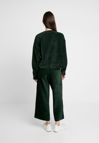 Donkergroen ribfluwelen sweatshirt en broek met wijde pijpen, met een losse pasvorm en zichtbare stikdetails, gecombineerd met witte sneakers.