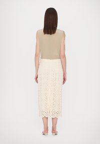 Une robe sans manches avec un haut beige et une jupe en dentelle crème aux motifs circulaires, arrivant au niveau du mollet. Les touches finales comprennent une fermeture éclair au dos.