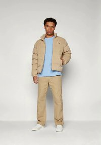 Μπεζ μπουφάν με υφή, ανοιχτό μπλε μπλουζάκι, μπεζ παντελόνι και λευκά αθλητικά παπούτσια; το μοντέλο στέκεται σε μια ουδέτερη τοποθεσία.