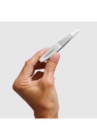 Mano che tiene una pinzetta Tweezerman in acciaio inox con impugnatura testurizzata e punta angolata, su uno sfondo grigio chiaro uniforme.