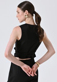 Top senza maniche nero con un design aderente e lunghezza cropped, rifinito con un orlo arricciato. Il modello ha i capelli lisci e indossa grandi orecchini quadrati dorati.