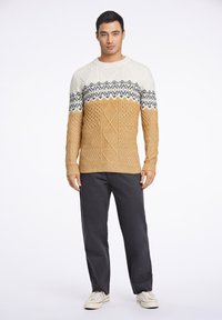 Stickad tröja med blockfärgad design i kräm och senap, med texturerade mönster och svarta detaljer, kombinerad med mörka byxor och sneakers.