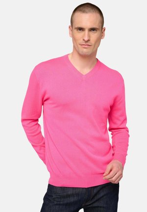 Mann mit kurzen Haaren, der einen leuchtend pinken V-Ausschnitt-Pullover und dunkle Jeans trägt, posiert mit einer Hand hinten verschränkt und der anderen in der Nähe der Taille.