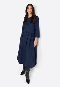 Navy blue kaksiosainen asu, johon kuuluu pitkähihaista puseroa ja ryhmäkäärmeitä, joita korostavat polven ylle ulottuvat mustat korkokengät. Sileä pinta.
