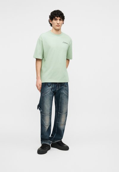 Jeune homme aux cheveux bouclés portant un t-shirt vert clair oversized et un jean bleu foncé ample avec des baskets noires, debout devant un fond blanc.