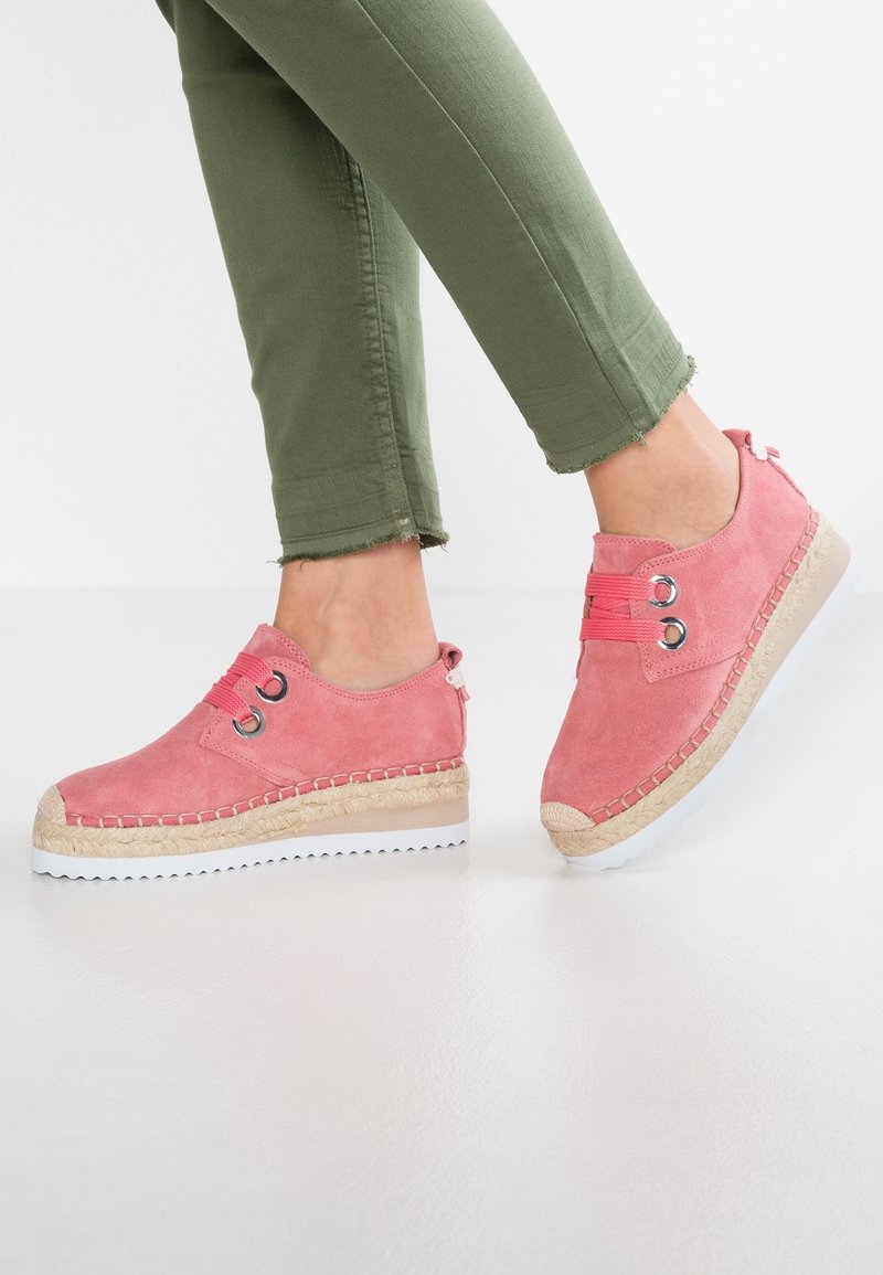Des pieds portant des chaussures décontractées en daim rose avec des semelles épaisses tissées beige et blanches, assorties à un pantalon vert coupé avec des ourlets effilochés.