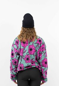 Fleece sweatshirt in mintgroen met een groot roze bloemenpatroon. Beschikt over lange mouwen en een relaxte pasvorm, gecombineerd met zwarte leggings en een beanie.