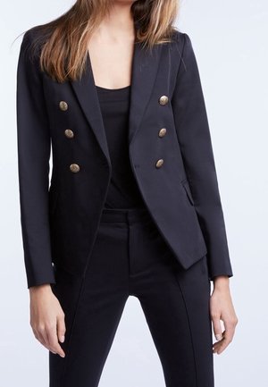 Vrouw draagt een zwart dubbelgeknopte blazer met gouden knopen en bijpassende zwarte broek, staand tegen een effen lichte achtergrond.