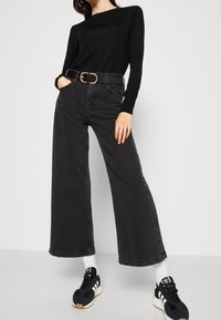Haut noir à manches longues associé à un jean en denim noir à jambes larges et des baskets noires. Le jean présente une taille haute et une ceinture texturée avec une boucle en or.