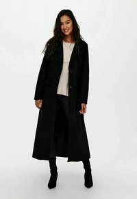Casaco longo preto com um design ajustado, apresentando uma frente com botões, bolsos laterais e uma textura suave. Usado sobre uma blusa leve com calças justas.
